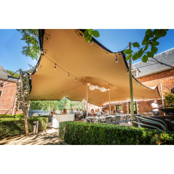 een stretchtent van 7,5x7,5 meter zit met houten palen vast in de grond. Er staan twee palen in het midden om de gehele tent omhoog te houden. de houten palen zijn vastgezet met touwen en haringen die in de grond zitten.