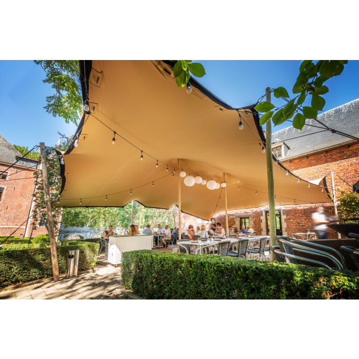 een stretchtent van 7,5x7,5 meter zit met houten palen vast in de grond. Er staan twee palen in het midden om de gehele tent omhoog te houden. de houten palen zijn vastgezet met touwen en haringen die in de grond zitten.