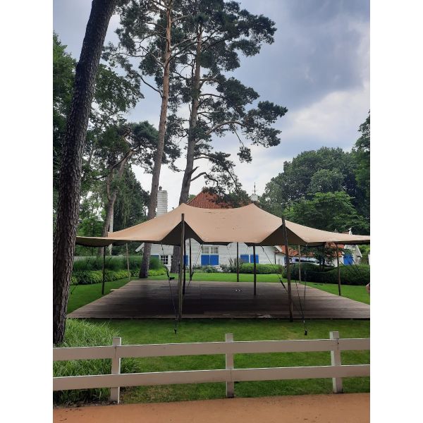 Een stretchtent van 10,5x10,5 meter zit met houten palen vast in de grond. Er staan twee palen in het midden om de gehele tent omhoog te houden. de houten palen zijn vastgezet met touwen en haringen die in de grond zitten.